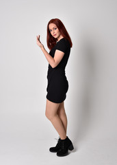 Portrait of a pretty girl with red hair wearing short black dress and boots, full length standing pose on a studio background.