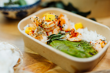 Vitamin-rich healthy dish with shrimps, rice and avocado in ecobox, delivered by a delivery service, ordered online, decorated with mussels and mango pieces on white wood, all in a Mediterranean style