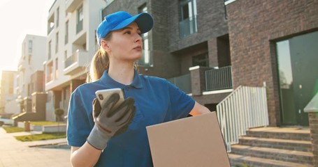 Caucasian young female courier in hat and gloves holding carton parcel and looking for address on smartphone. Pretty woman with phone smiling to camera outdoor when working as delivering worker.