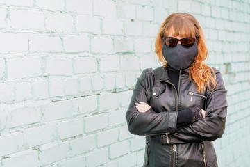 A young red-haired girl in sunglasses and a black medical mask walks the streets of the city during the coronavirus epidemic. Girl in black on a background of a brick wall.