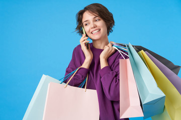 Fototapeta na wymiar Portrait of cheerful young lovely brown haired curly lady keeping shopping bags in raised hand and smiling positively while making call, isolated over blue background