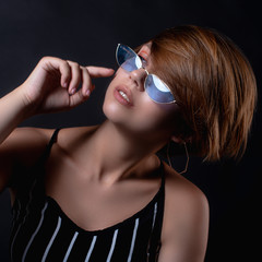 stylish brown-haired girl in blue sunglasses holds hand on chin looks to the left on black...