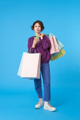 Puzzled young pretty short haired curly female keeping mobile phone in raised hand and looking pensively upwards, posing over blue background with shopping bags
