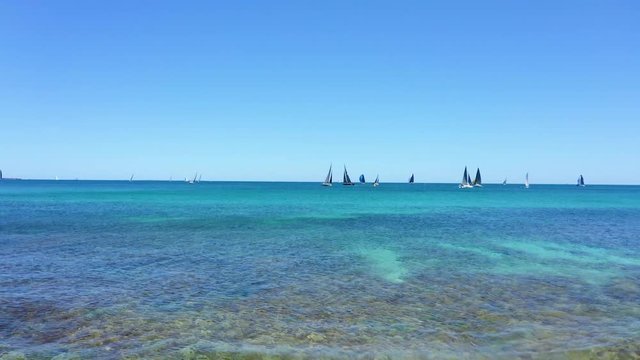 Low aerial fast drone across gorgeous turquoise water toward sailboat regatta
