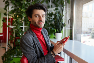 Portrait of young attractive brown haired bearded man holding smartphone in raised hand and looking gladly at camera with light smile, isolated over city cafe interior