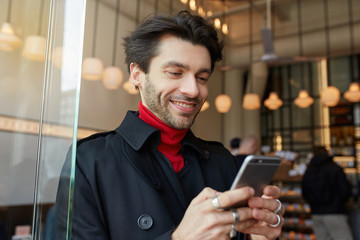 Portrait of young lovely brown haired unshaved male looking happily on screen of his phone and smiling cheerfully while standing over city cafe interior in trendy clothes
