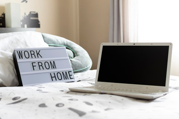 Zenithal picture of a light box with a message and a laptop with black screen on a bed in a bright bedroom.Work from home message.