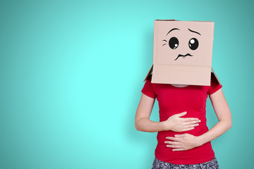 Person with cardboard box on its head and a grumpy face expression holding ist belly on turquoise background