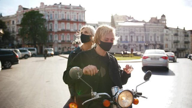 Portrait of happy young hipster couple riding a vintage scooter in city in protective masks during pandemia of Covid-19, slow motion