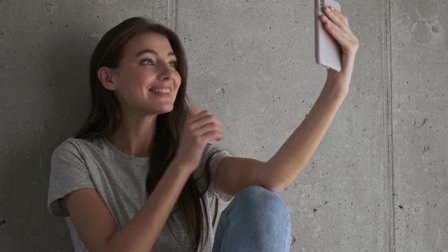 Pleased pretty brunette woman posing and making selfie on smartphone while sitting near the wall at home