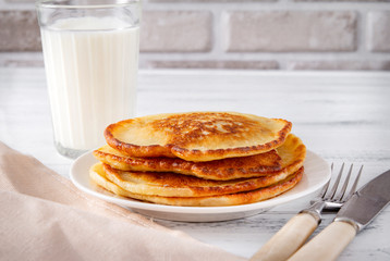 pancakes on a white plate, a glass of milk on the table, a fork, knife, napkin on a white wooden background