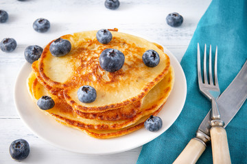 pancakes with fresh blueberries on a plate, fork, knife on a blue napkin on a white wooden , top view,