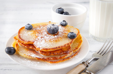 pancakes with fresh blueberries with powdered sugar on a plate, a glass of milk, a fork, a knife on a white wooden background,