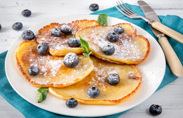 fried pancakes with fresh blueberries with powdered sugar and mint leaves on a plate, knife and fork on a blue napkin, on a white