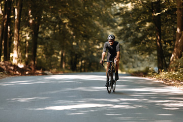 Fototapeta na wymiar Full length portrait of bearded sportsman in black clothes, helmet and eyeglasses doing cycling workout on paved road among beautiful nature. Concept of active lifestyle.