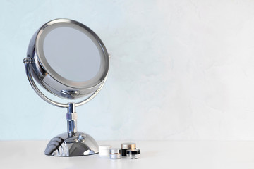 Chromed steel round magnifying mirror on handle set up on the white table with some jars with creams and cosmetics. Cold light blue background, copy space for any text or design work.