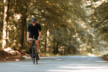 Active bearded man riding bike among green forest in black protective helmet and sportswear. Mature cyclist in eyeglasses training on fresh air.