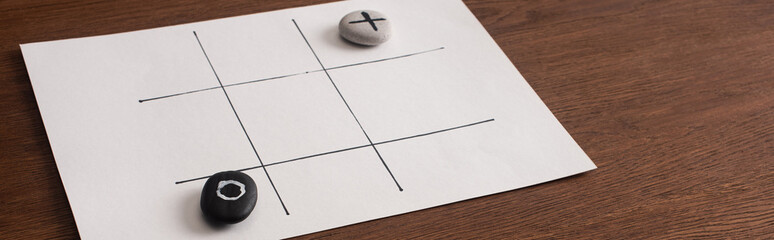 panoramic shot of tic tac toe game on white paper with pebbles marked with naught and cross on wooden surface