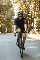 Fototapeta na wymiar Front view of bearded man with athletic body shape dressed in sport clothing and black helmet biking in forest. Sporty person in mirrored eyeglasses enjoying summer nature during workout.
