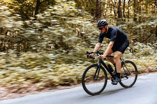 Athletic bearded man in glasses and black helmet riding bike among green forest, motion blur. Cyclist in activewear doing sport outdoors. Activity concept.