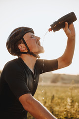 Side view of exhausted bearded sportsman in black helmet pouring water from sport bottle on his...