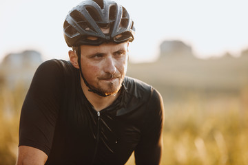 Portrait of sporty man in black helmet and cycling clothes being wet because of hard training and riding long distance. Professional bicyclist relaxing and gaining strength with blur background behind - obrazy, fototapety, plakaty
