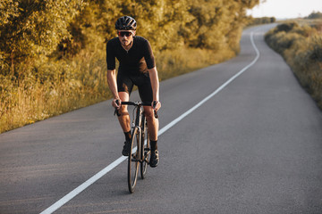Strong bearded athlete in cycling clothes riding bike on paved road with green trees around. Active...