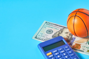 Basketball ball, calculator and banknote of 100 dollars on blue background. Purchasing sport...