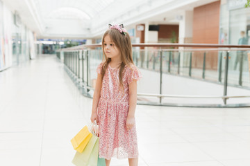 Portrait of a little happy girl in the mall. A smiling laughing girl in a pink dress with a cute rim with ears and with multi-colored bags in her hands is walking around the mall, looking at the