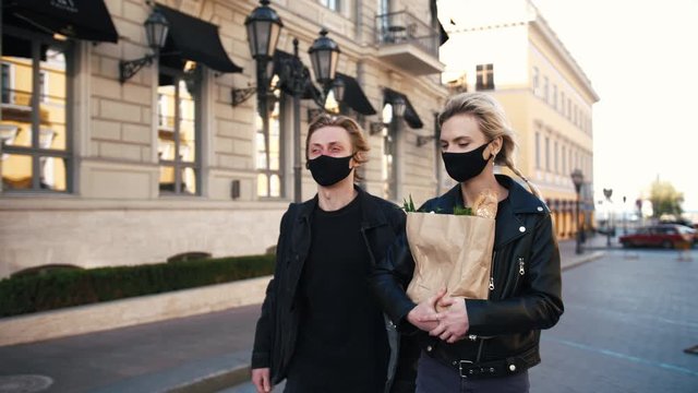 Portrait of happy young couple in protective masks walking outdoors in old city center with grocery paper bag during sunset