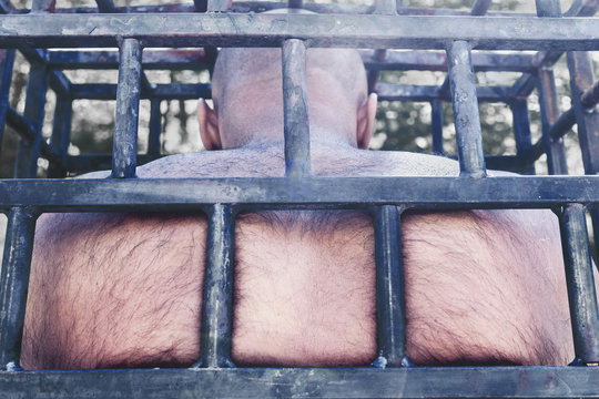 The bald man in the cage pressed his back against the iron bars. concept of prison and imprisonment