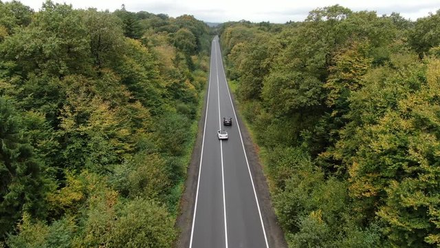Overtaking cars. Aerial view flying over road with cars moving, that through lane green forest. Cars driving along the forest road. Cars driving through forest. Drone shot from above.