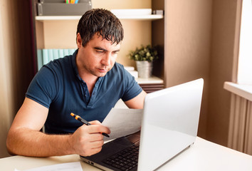 A serious man with dark hair at home at his laptop. Remote work. A man at a Desk with documents works online.