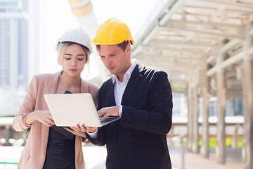 Male and female engineers wearing protective caps are looking at building construction plans from a notebook in the outdoors. With a backdrop to the building structure