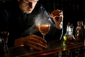 Young male bartender sprinkle to alcoholic cocktail with piece of citrus zest