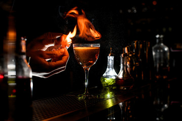 Close-up bartender sprinkle to alcoholic cocktail with piece of citrus zest and make fire over it