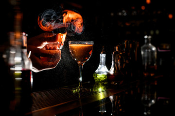 bartender's hand sprinkle to alcoholic cocktail with piece of citrus zest and make fire