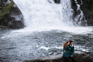 Fototapeta na wymiar A couple of lovers in green raincoats, sitting on a rock, against the backdrop of a waterfall