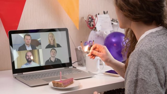 Woman celebrating her birthday through video call virtual party with friends. Lits and blows out candle. Authentic decorated home workplace. Handheld shot with gimbal. Coronavirus outbreak 2020.