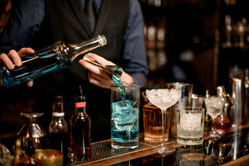 bartender pours blue drink from jigger into glass with pieces of ice