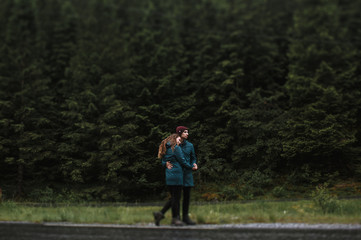 Obraz premium Lovers in green raincoats look at each other, standing next to the highway against the background of a green forest