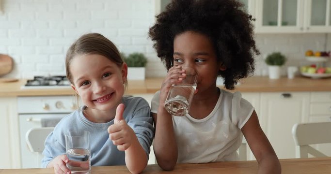 Two funny adorable mixed race children girls holding glasses drinking tasty clean pure mineral water showing thumbs up looking at camera, recommend daily children healthcare morning hydration concept.