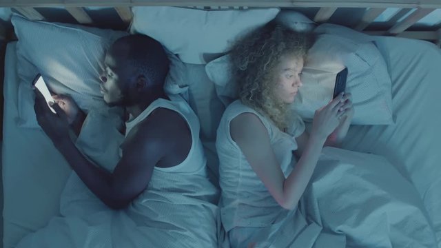 Directly above view of African man and mixed-race woman lying back to back in bed with telephones in their hands