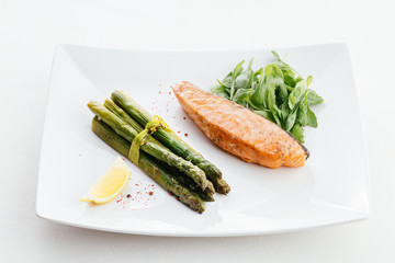 Red fish steak with different vegetables. Green asparagus, lemon and herbs. On a white plate.