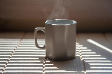 a mug of hot tea on a wooden table. shade from the window blinds in the morning. steam from a mug of tea