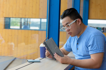 Young overweight Asian nerd man reading book at the coffee shop