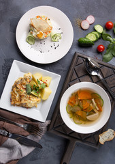 Complex dinner. Vegetable soup, salad Olivier and potatoes with fish on white plates on a dark gray background. Fresh vegetables and appliances. Top view, flatlay. Background image, copy space