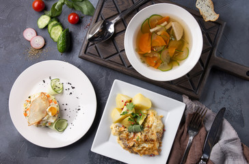 Complex dinner. Vegetable soup, salad Olivier and potatoes with fish on white plates on a dark gray background. Fresh vegetables and appliances. Top view, flatlay. Background image, copy space