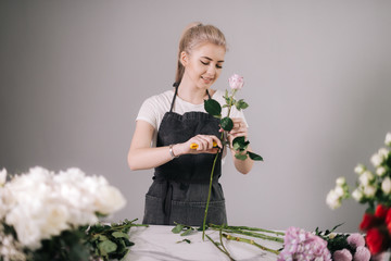 Beautiful young woman florist wearing apron working with fresh rose at the table on white...