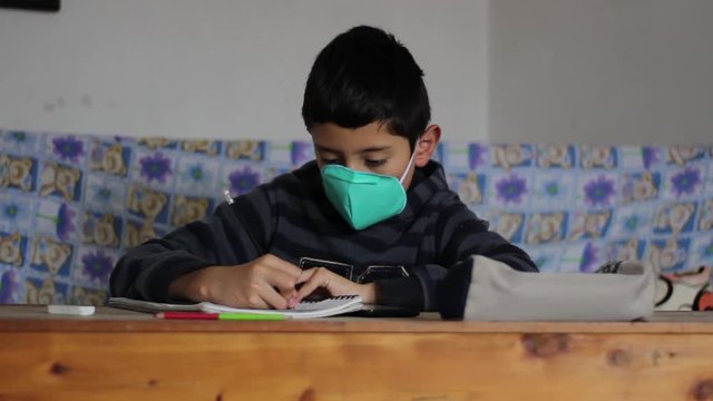 Ni&ntilde;o en cuarentena escribiendo para su tarea de la escuela. Tienen puesto su tapabocas. 